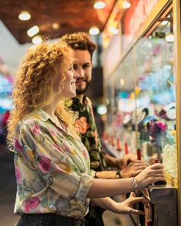 Uśmiechnięta para bawi się przy automacie z chwytakiem w salonie gier. Są ubrani w jasne kolory i wyglądają na radosnych, a rozmyte światła tworzą żywą, zabawną atmosferę.