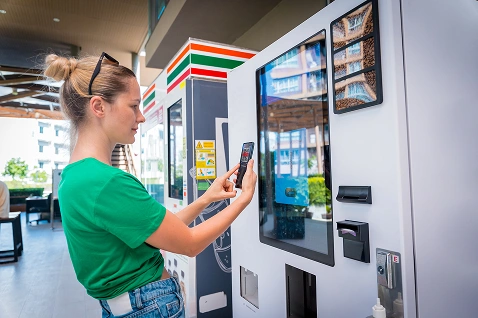 Kobieta w zielonej koszuli korzysta ze swojego smartfona, aby obsługiwać nowoczesny automat sprzedający. Akcja rozgrywa się w jasnej, otwartej przestrzeni o swobodnej atmosferze.